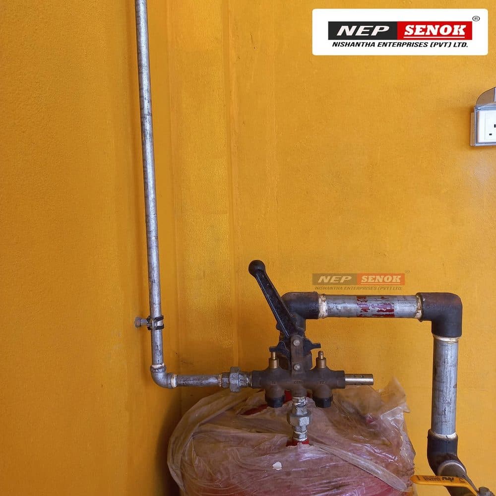 A brass control-valve manifold with a black lever handle, connected to galvanised air-line pipework, mounted above a red tank wrapped in protective film, against an ochre-painted wall.