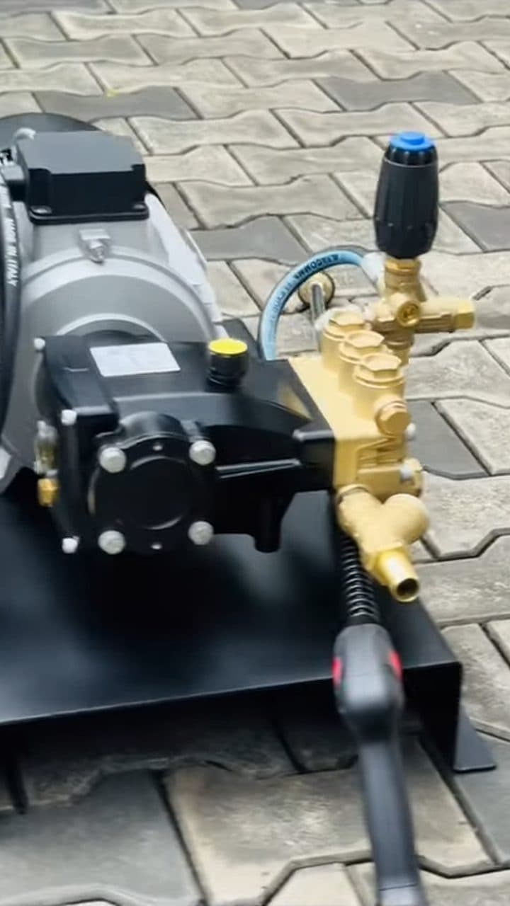 A grey electric motor bolted to a brass pump head, with a black pressure-adjustment knob, a coiled hose and a trigger spray gun, mounted on a black steel skid on paved ground.