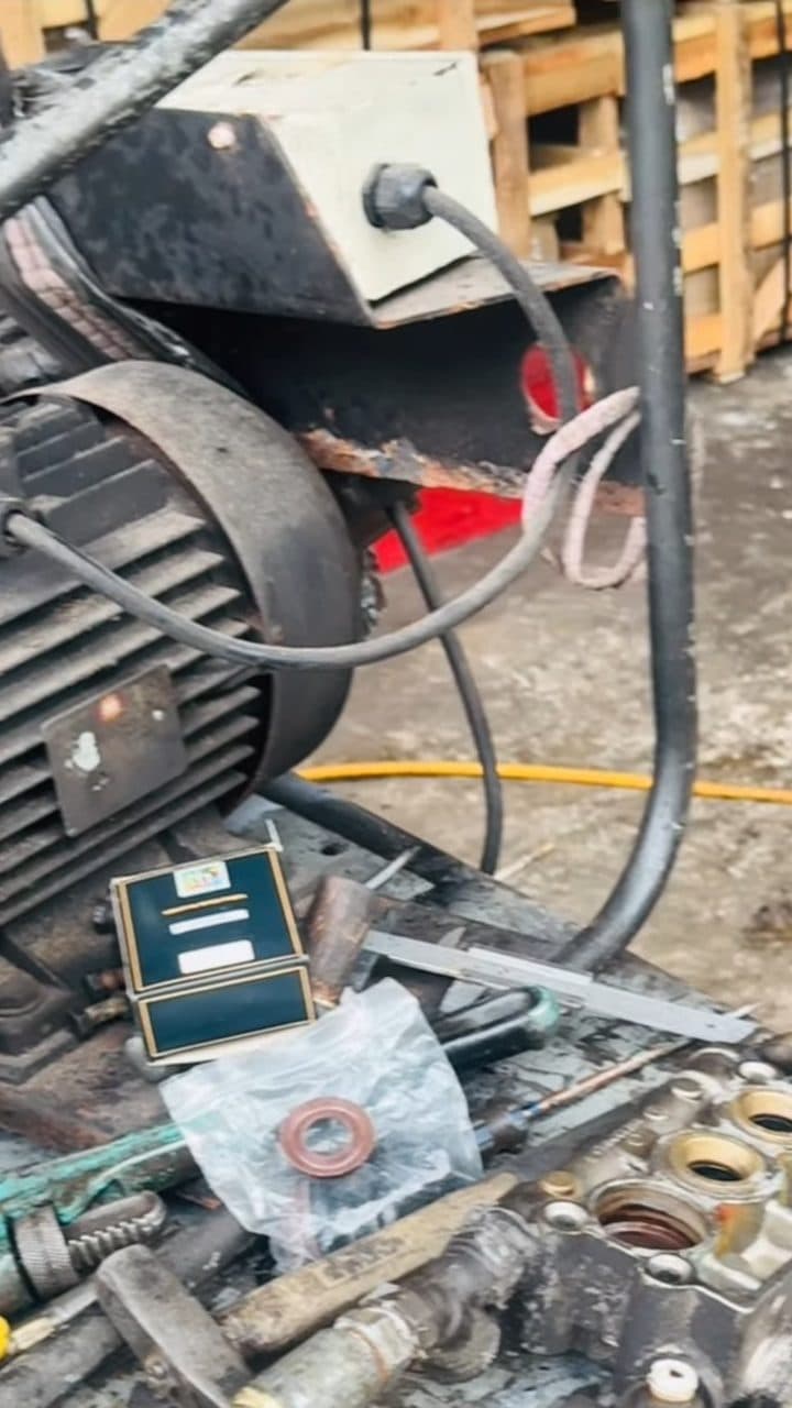 A close-up of an electric motor with a V-belt pulley beside a stripped-down pump housing, with tools, a vernier caliper and an oil seal in a plastic bag laid out on the workshop floor, stacked wooden pallets behind.