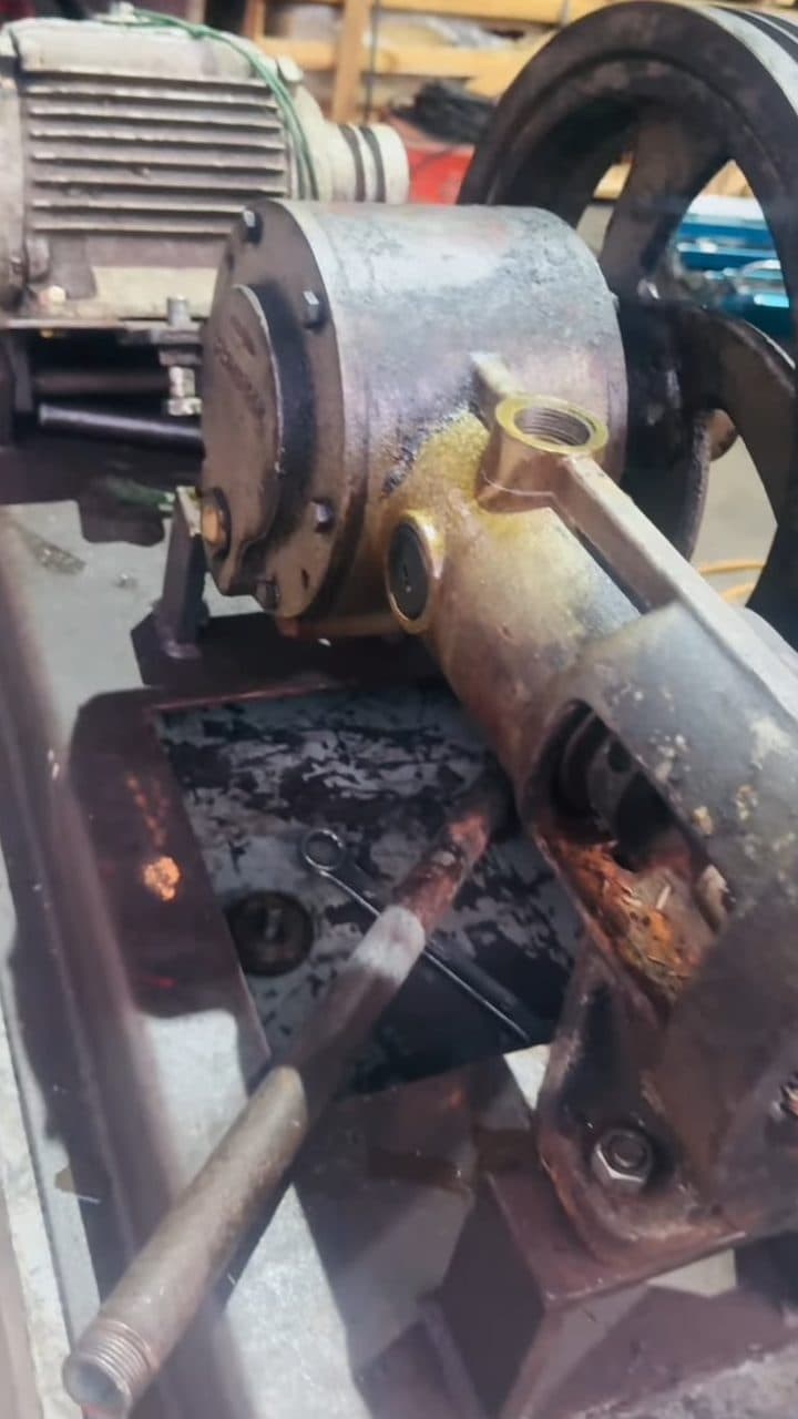 A close-up of a single-plunger pump's cylinder head bolted to an electric motor, its drive pulley and flywheel behind it, sitting over an oil-catch tray on a rusted steel stand.