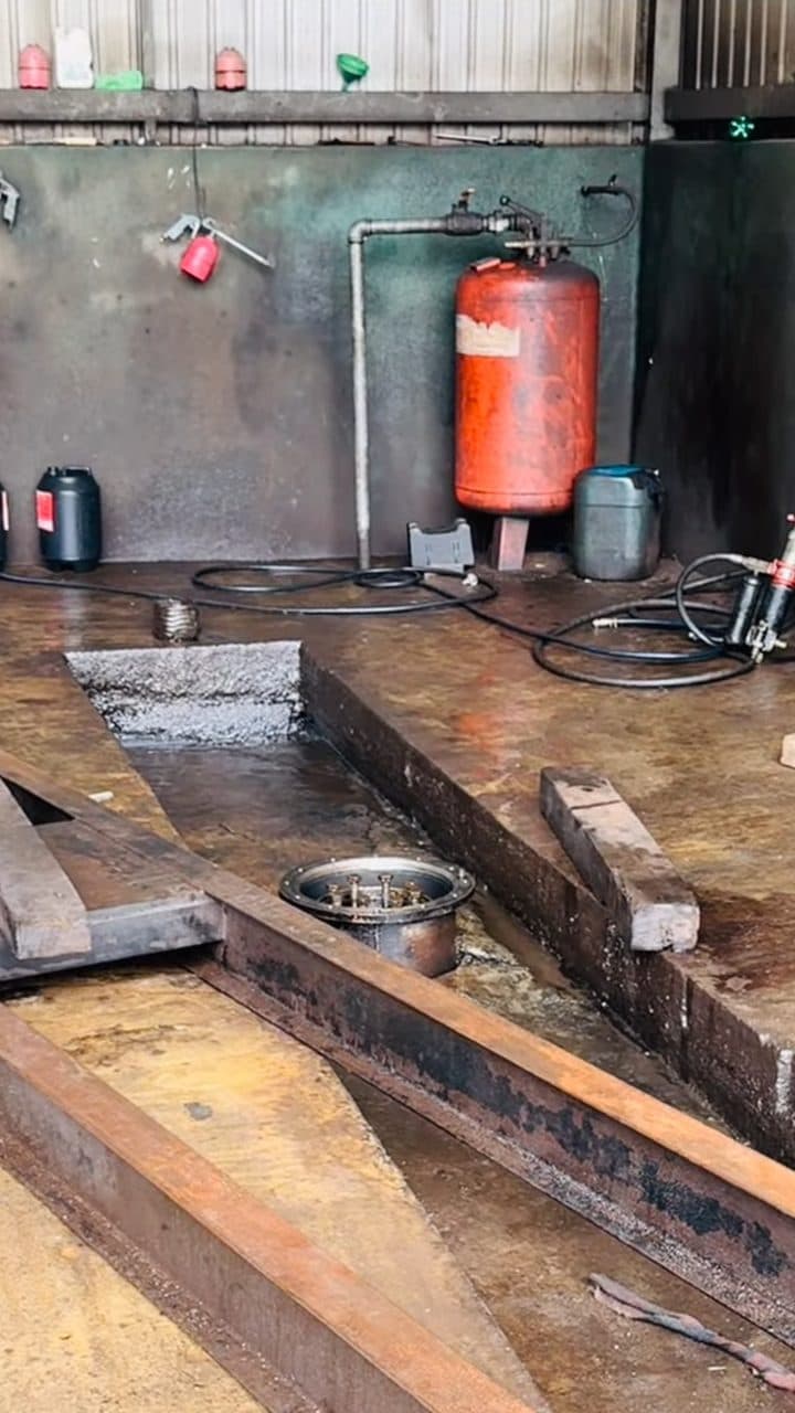 A workshop pit shot from above: a bolted flange plate set in the floor of an in-ground pit, a red air receiver tank on the wall behind it, coiled hoses and a wash gun nearby, and timber baulks laid across the wet concrete.